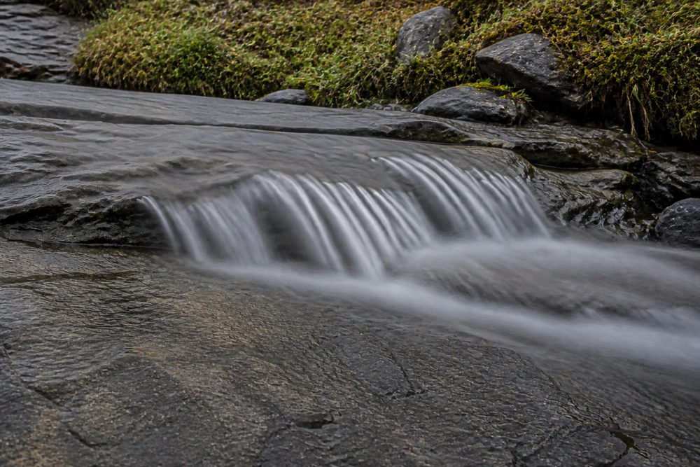 Illustration générée : le même ruisseau dont l'eau est lissée en voile, tel que le rendent f/16, 2 s, ISO 100 à 24 mm